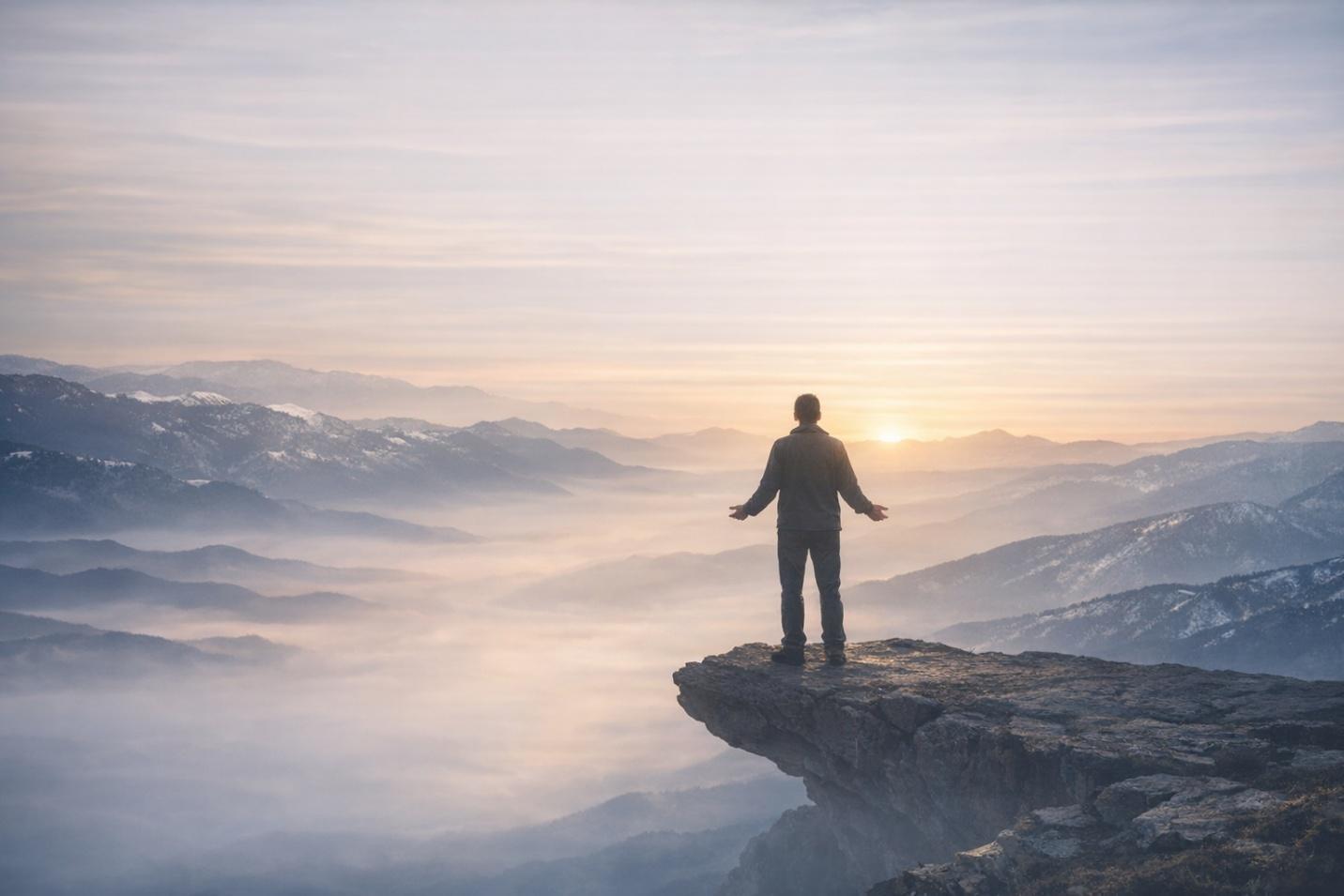 A figure standing on a mountain cliff at sunrise above misty valleys.